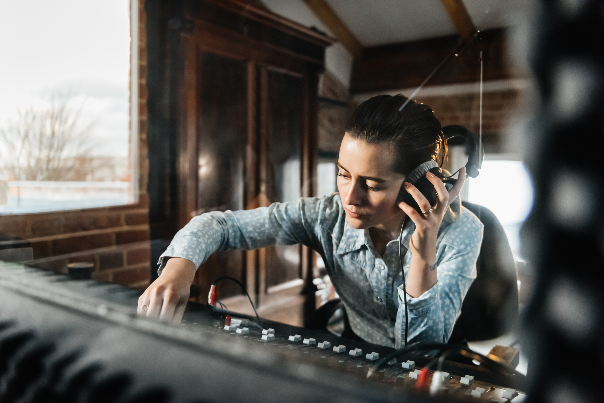 Jeune femme devant une table de mixage portant un casque sur son oreille gauche et mixant de la main droite