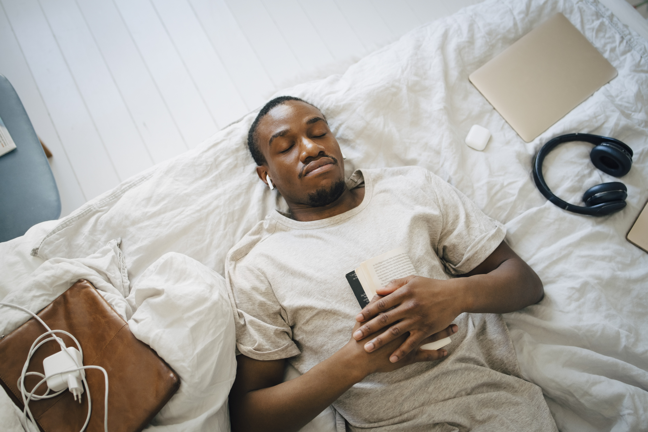 Jeune homme allongé sur son lit. Il écoute une fiction audio avec ses Airpods et tient un livre contre sa poitrine. Divers objets électroniques comme un chargeur ou un casque sont posés sur son lit.