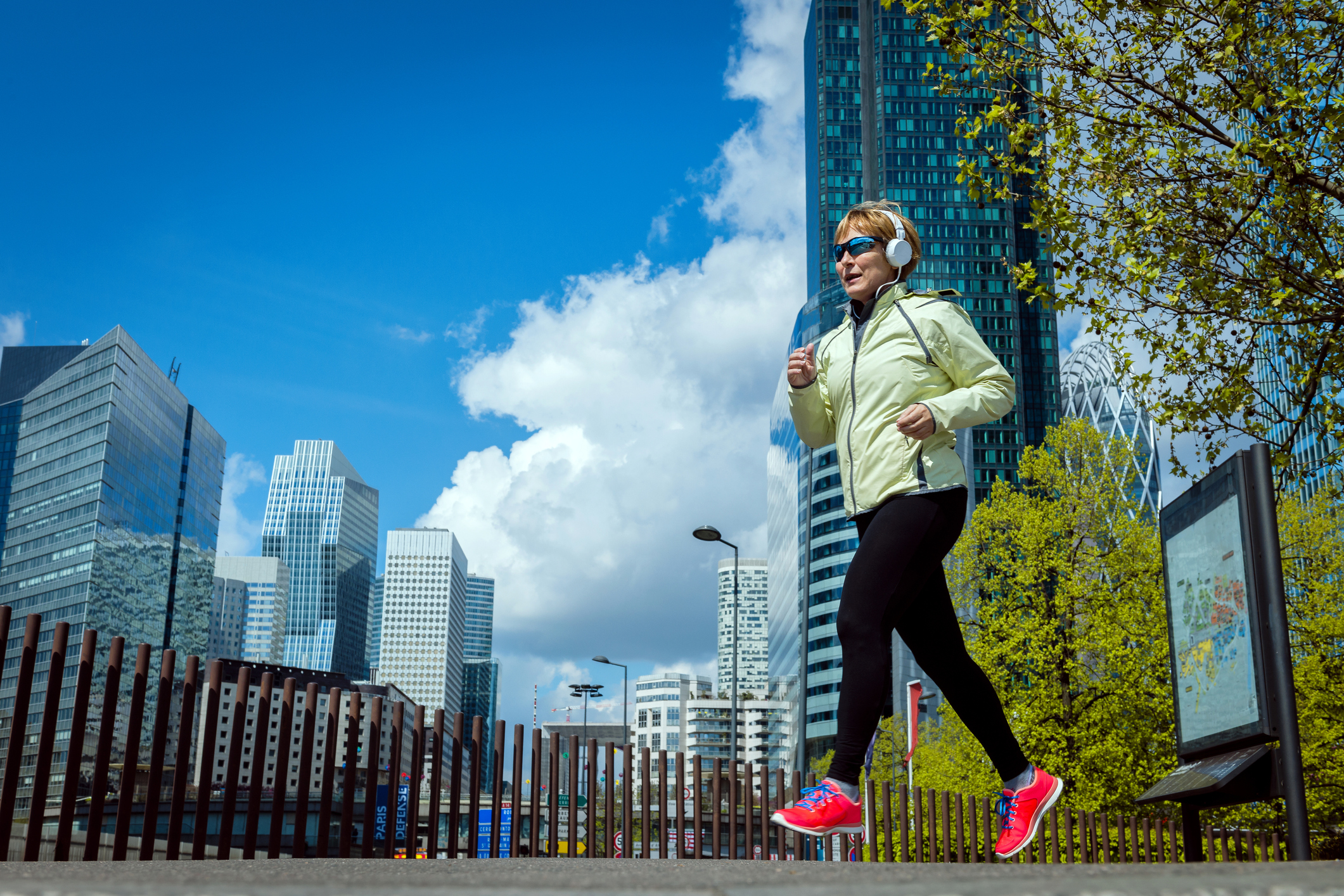 Femme âgée faisant du sport (course) en écoutant de la musique avec des écouteurs et portant des lunettes de soleil à La Défense à Paris.