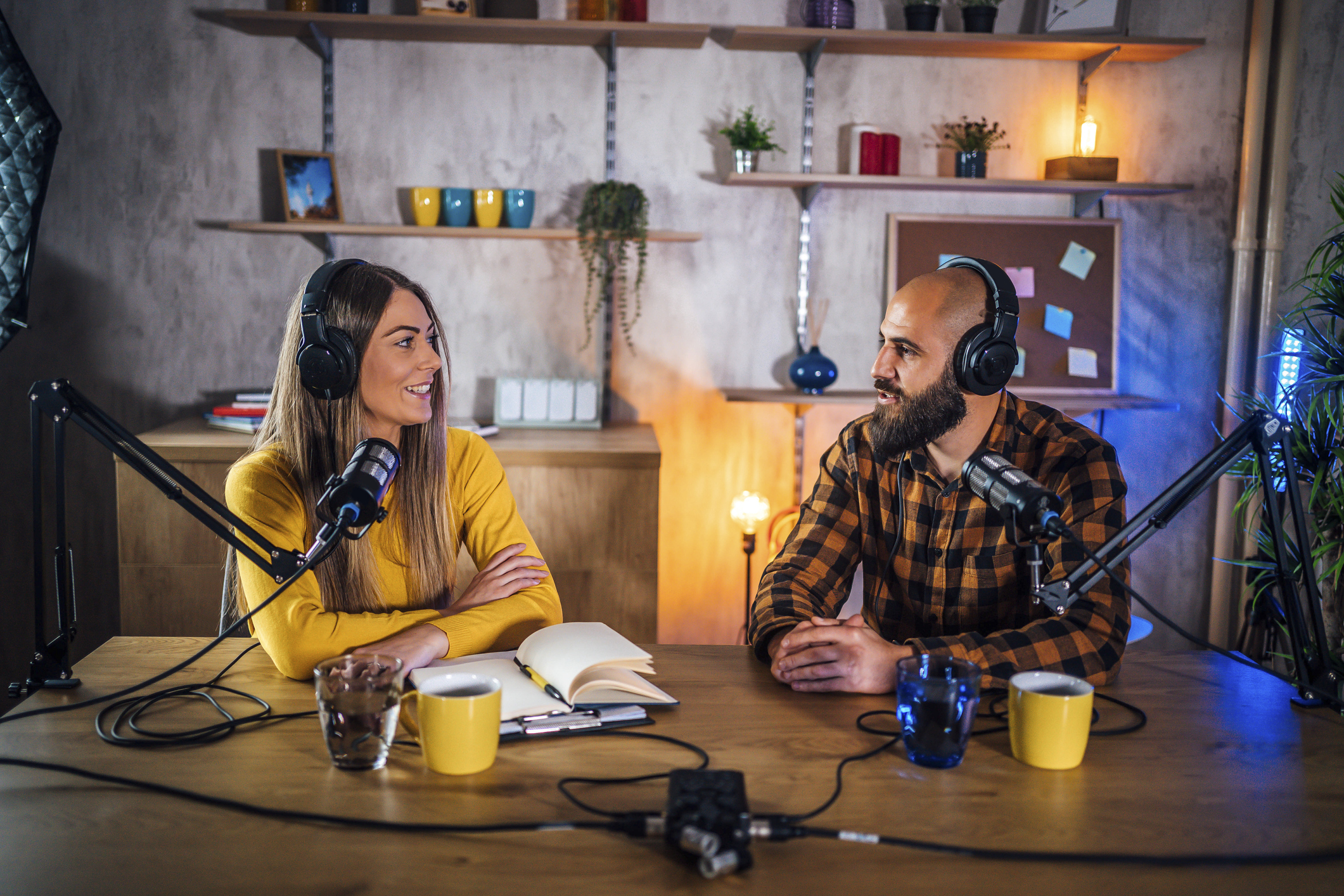 Une jeune femme à gauche et un jeune homme à droite enregistrent un podcast grâce à des micros professionels. La jeune femme blonde porte un pull jaune et le jeune homme chauve et barbu porte une chemise à carreaux orange et noire. Elle a un carnet devant elle, et ils ont chacun une tasse jaune et un verre d'eau posés sur la table.