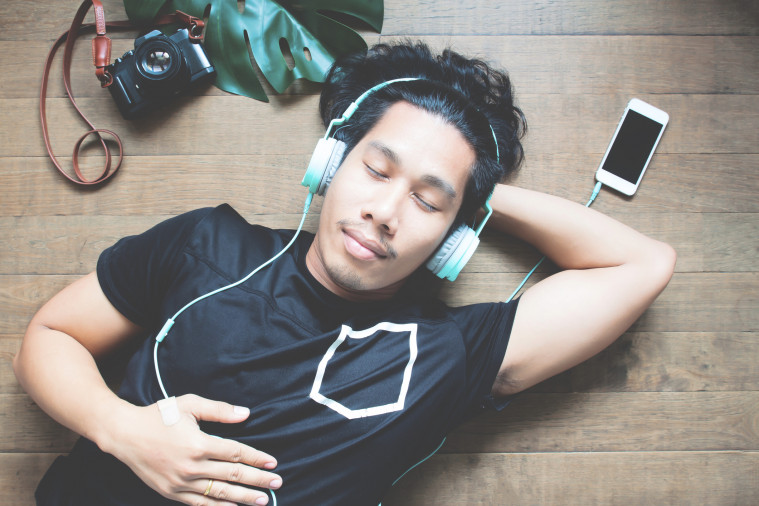 Un homme Thaïlandais allongé au sol, la main sur la poitrine, porte un casque bleu branché sur un smartphone posé sur le parquet. A gauche de son visage se trouvent un appareil photo et une feuille de Monstera.
