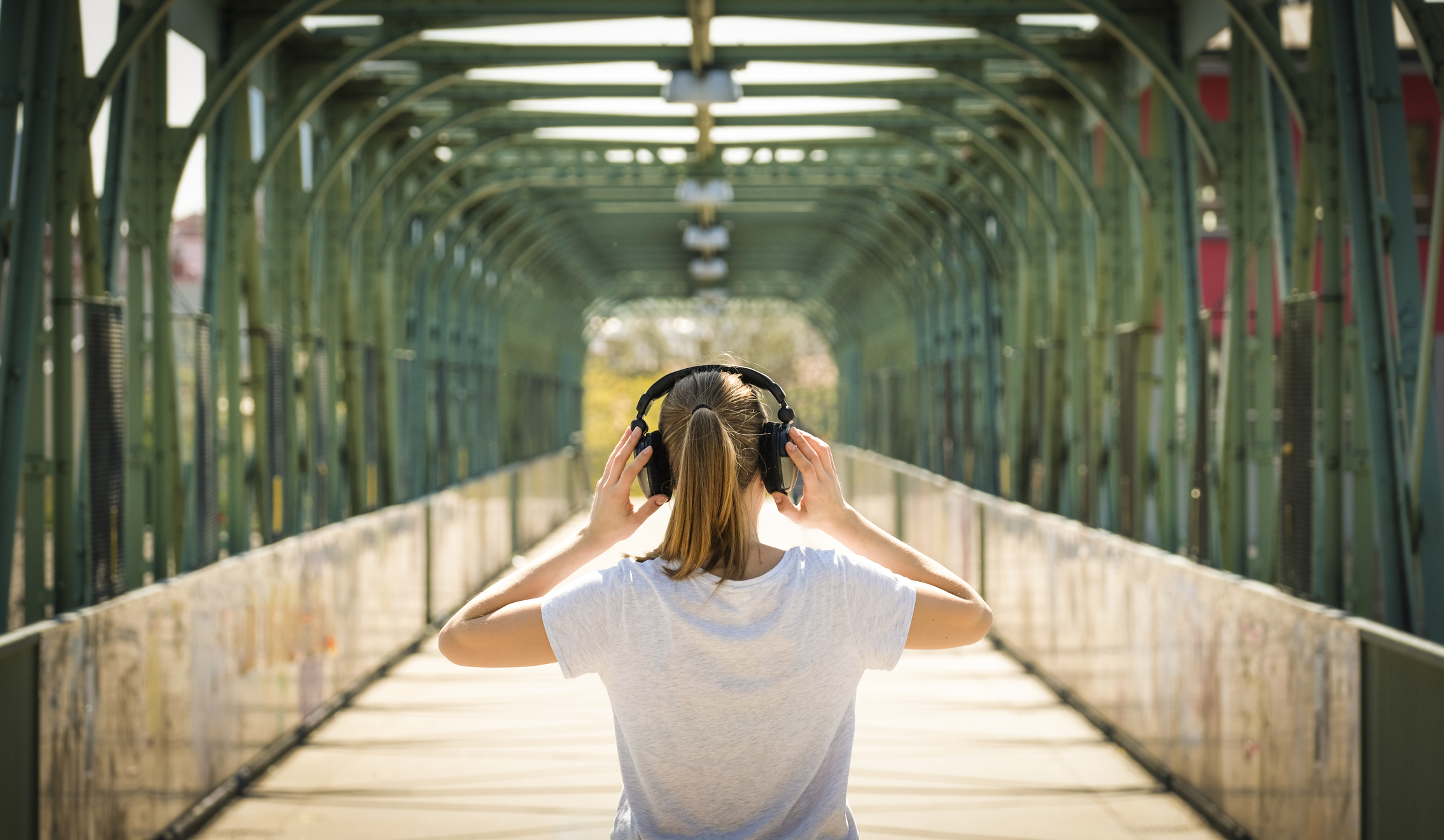 Une jeune femme vue de dos portant un casque de musique et le tenant à deux mains. Elle se tient sur un pont ensoleillé.